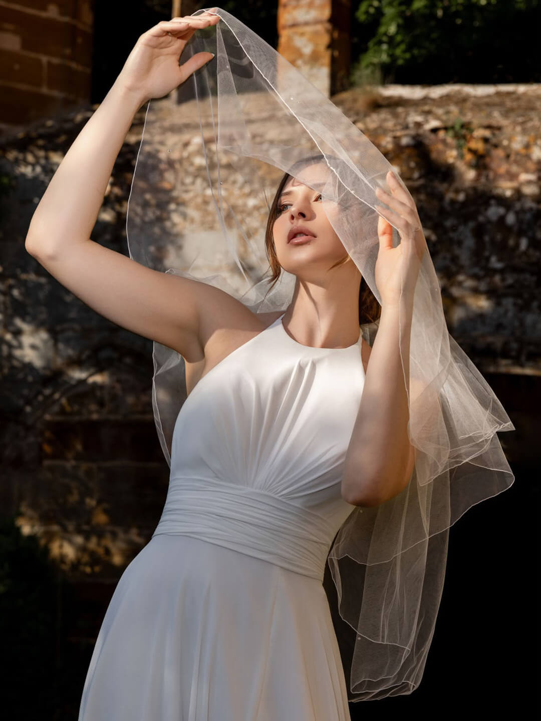 Braut in einem schlichten weißen Brautkleid hält ihren Schleier mit beiden Händen in die Höhe. Das Porträt entstand im warmen Sonnenlicht vor einer Steinmauer.