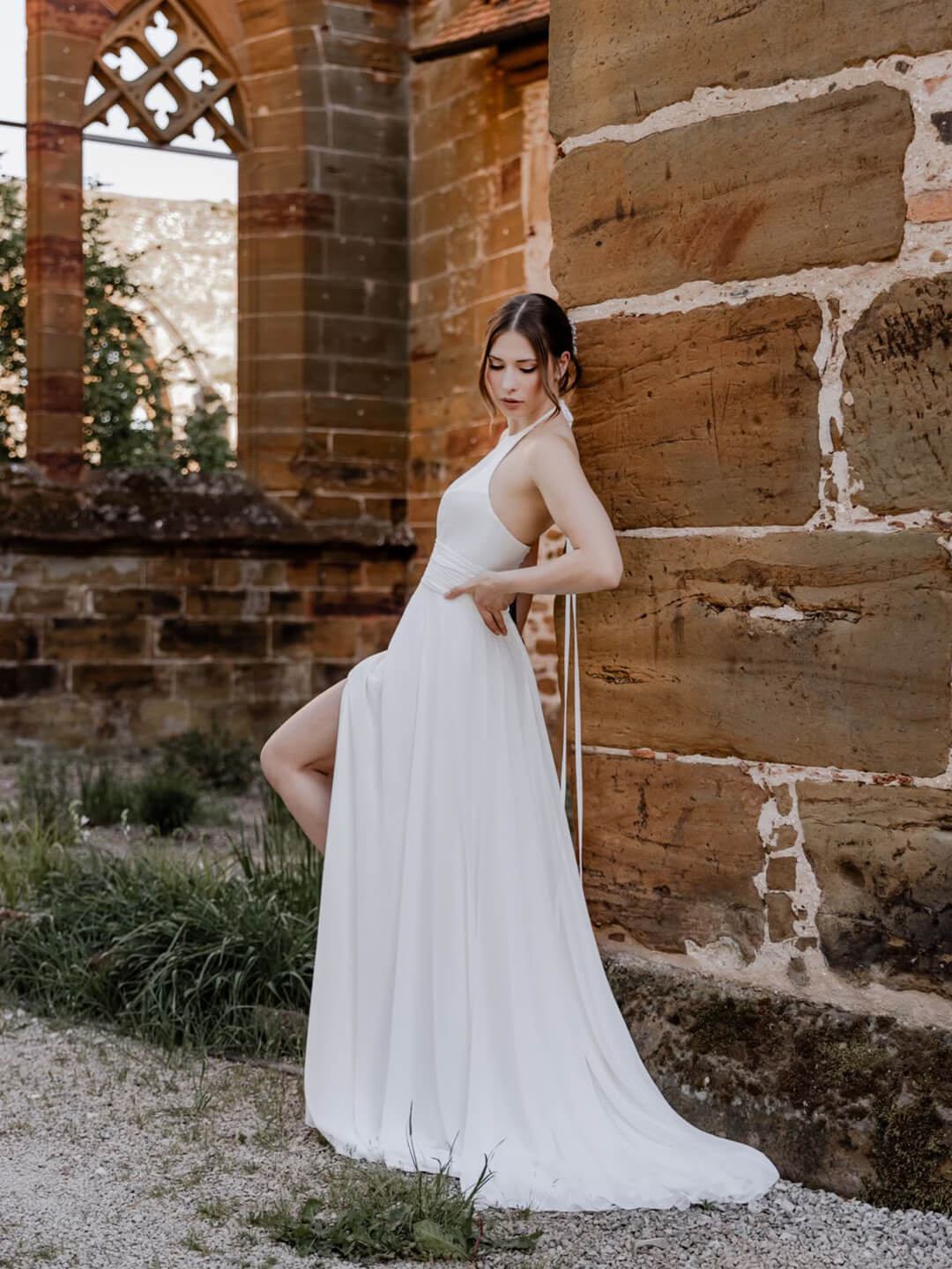 Braut in einem schlichten weißen Brautkleid mit Neckholder-Ausschnitt lehnt an einer historischen Sandsteinmauer. Das bodenlange Kleid fällt fließend und hat einen seitlichen Beinschlitz. Im Hintergrund sind die Bögen einer alten Kirchenruine zu sehen.